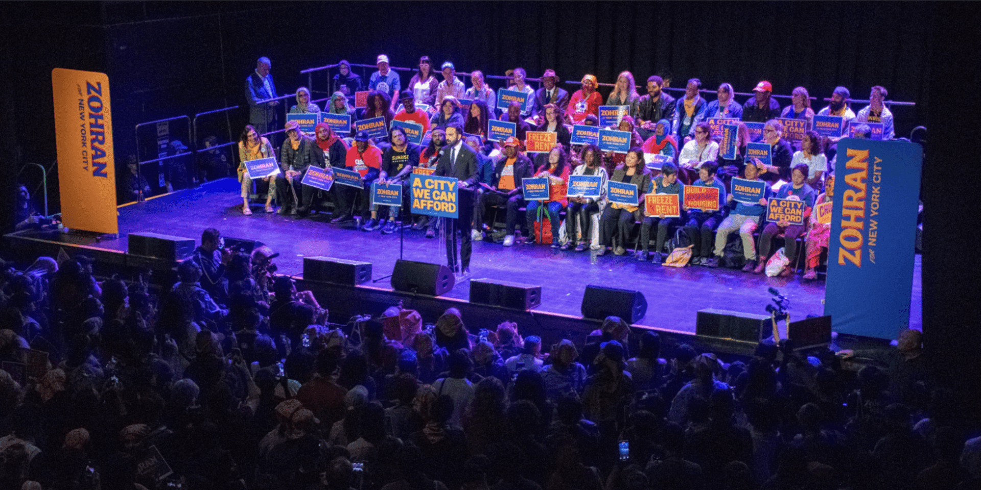 Zohran Mamdani stands at a podium in front of a crowd of supporters who hold signs.